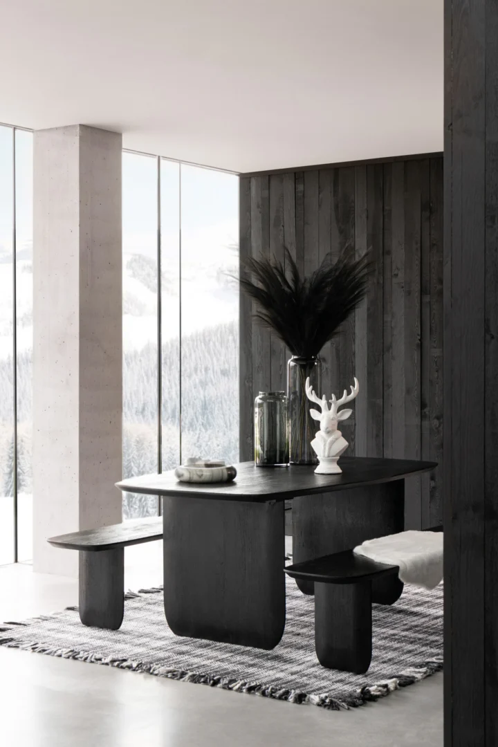 Een moderne eethoek met de Eettafel Acaciahout Zwart met bijpassende banken, een gestreept tapijt, grote ramen met uitzicht op besneeuwde bergen en minimalistisch decor zoals een vaas met zwart pampagras en een sculptuur van een witte hertenkop.