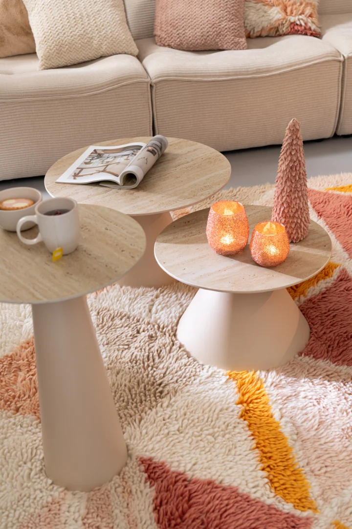 Drie ronde Bijzettafels (beige, roest, keramiek, 40 x 63 cm) zitten op een vloerkleed met structuur; op de ene staan twee kopjes en een tijdschrift, op de andere staan twee brandende kaarsen en een roze boom. Een beige bank met kussens staat op de achtergrond.