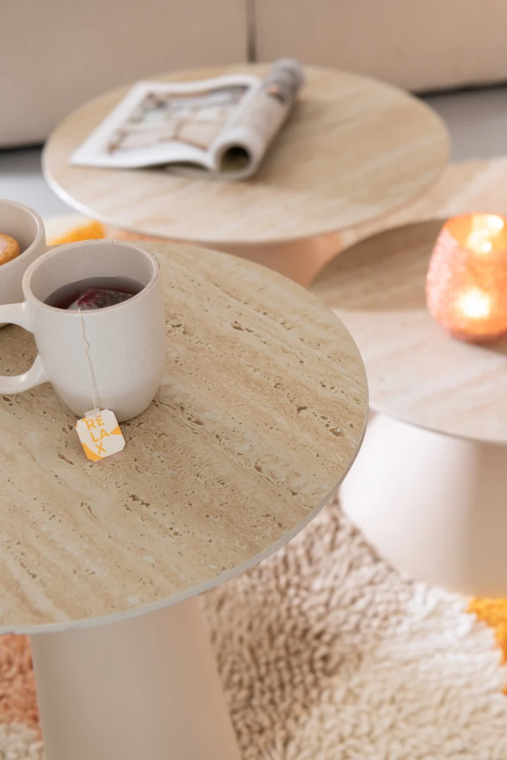 Een close-up van de Bijzettafel Beige, Roest 40 x 63 CM, een lichte salontafel van keramiek, toont een theemok, gebak en een kaars. Op de achtergrond ligt een tapijt met structuur en een beige bank. Op een andere tafel ligt een open tijdschrift.