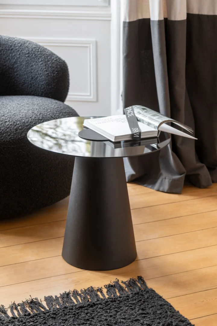 De Bijzettafel Zwart 60 x 50 CM Vierkant, gemaakt van metaal, heeft twee opengeslagen boeken. Het staat op een houten vloer naast een stoel met donkere textuur, met een zwart vloerkleed en grijze gordijnen in de buurt. Natuurlijk licht valt naar binnen door het raam op de achtergrond.