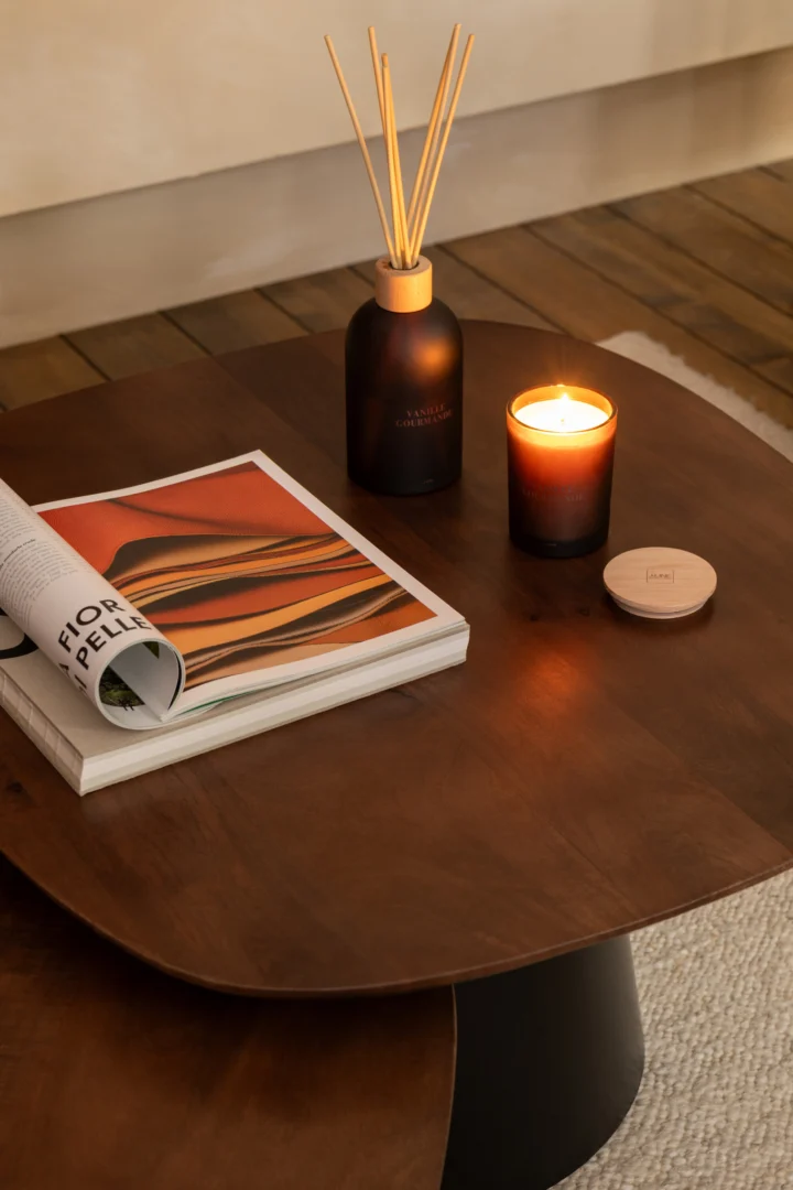 A Bijzettafel Zwart, Bruin 74 x 43 CM van vierkant mangohout, met een brandende kaars, een zwarte geurdiffuser, een gesloten pot en een open tijdschrift met kleurrijke abstracte kunst op de cover.