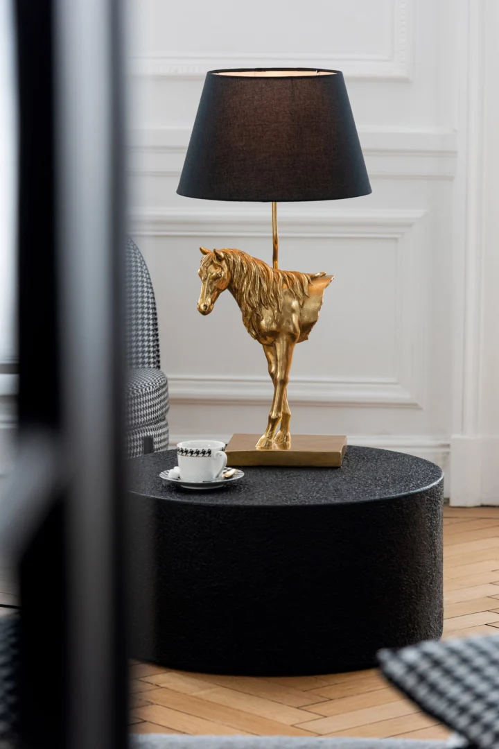 Een tafellamp met een gouden paardenvoet en zwarte kap staat op Salontafel Zwart 60 x 60 CM Vierkant Mdf. Een wit theekopje en schoteltje met een zwart patroon staan ook op de tafel, tegen een achtergrond van wit gelambriseerde muren.