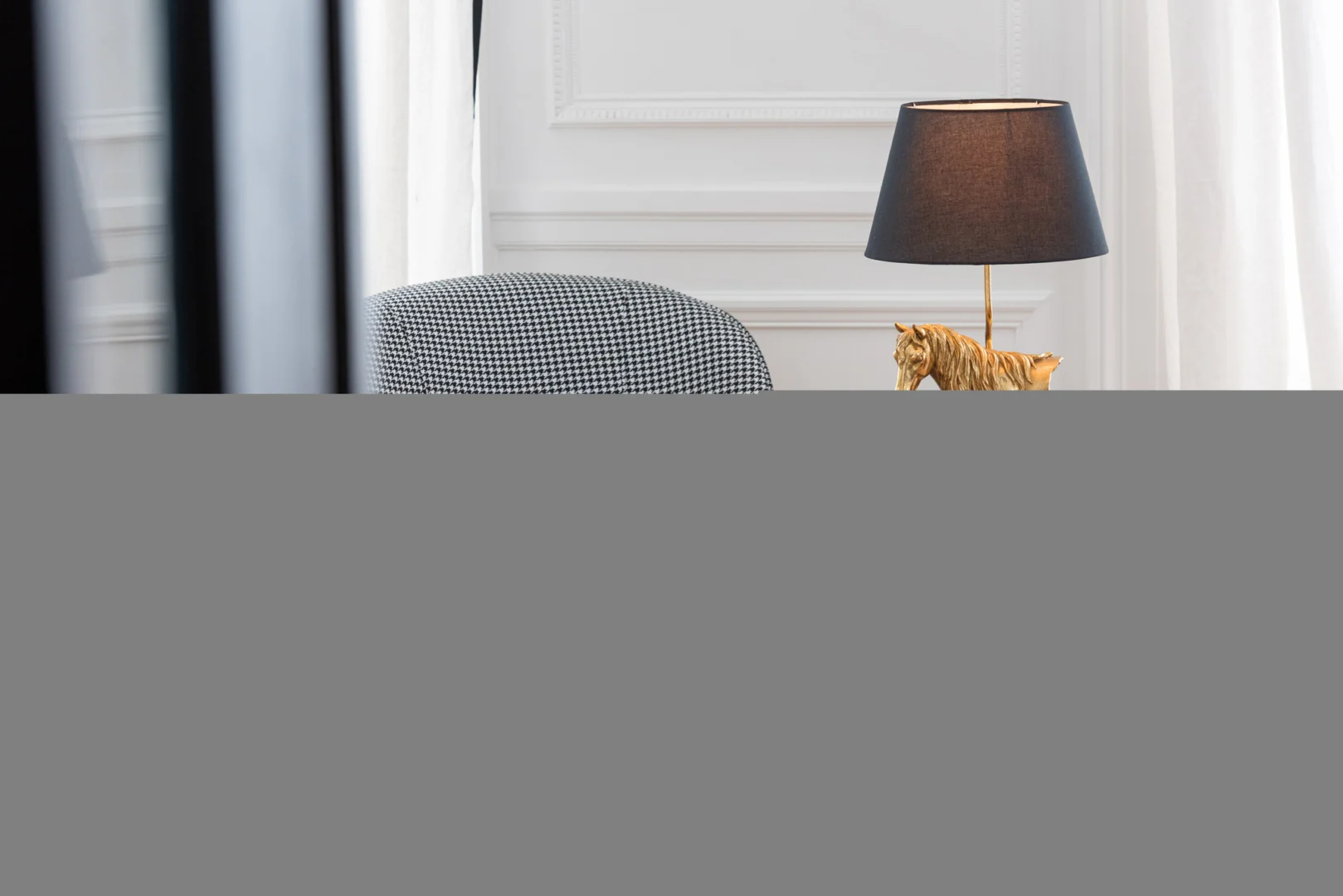 Een modern interieur met de Salontafel Zwart 60 x 60 CM Vierkant Mdf, een zwart-wit geblokte stoel, een tafellamp met een donkere kap en een gouden paardenfiguur, tegen muren met witte panelen en transparante gordijnen.