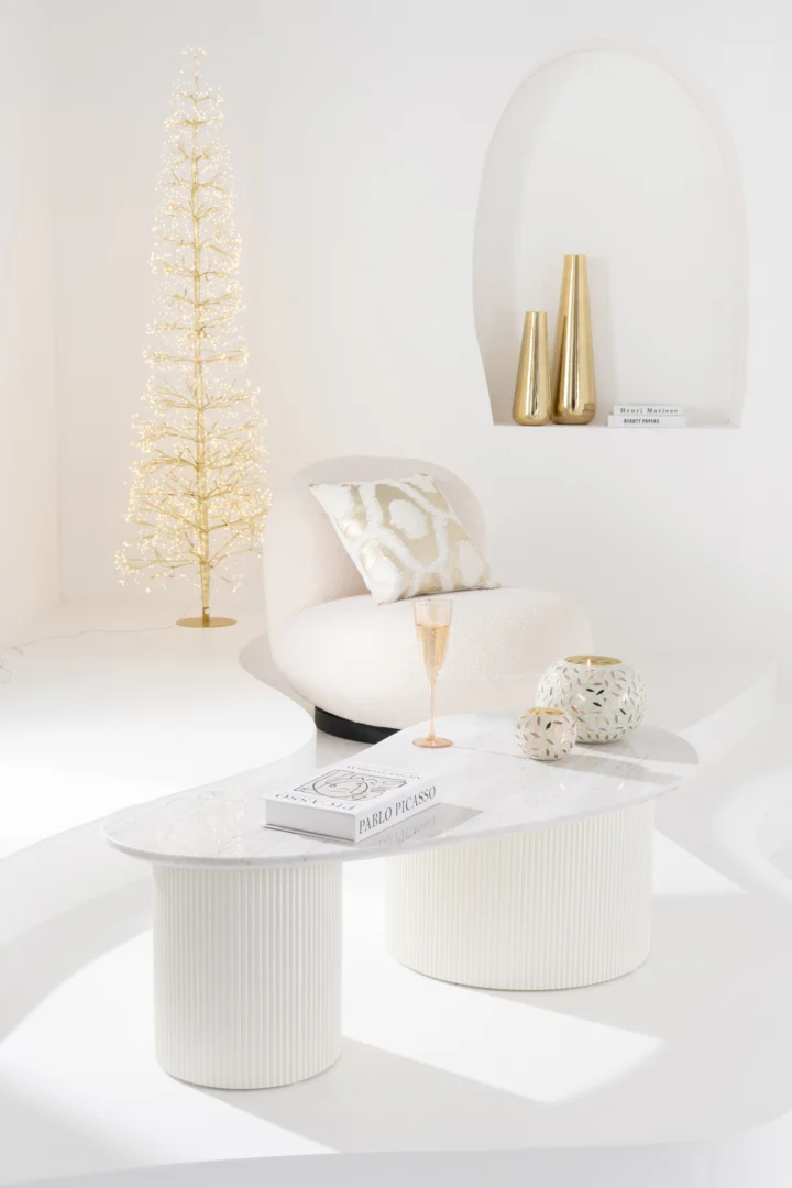 Minimalistische witte woonkamer met een moderne witte stoel, Salontafel Roest in wit marmer, gouden vaasdecoratie, oplichtende gouden kerstboom, Picassoboek, kussen met patroon en een champagneglas op tafel.