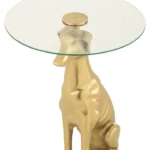 De Bijzettafel Transparant 46 x 58 CM is een ronde bijzettafel met een transparant glazen blad en een aluminium onderstel in de vorm van een goudkleurige zittende hond, die lijkt op een windhond.