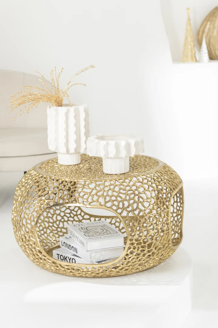 De Salontafel Goud 64 CM Aluminium - een ronde salontafel met een ingewikkeld patroon in goud - staat in een minimale, heldere omgeving. Bovenop staan twee witte vazen en een wit decorobject; binnenin staan boeken met het label LONDON en TOKYO.
