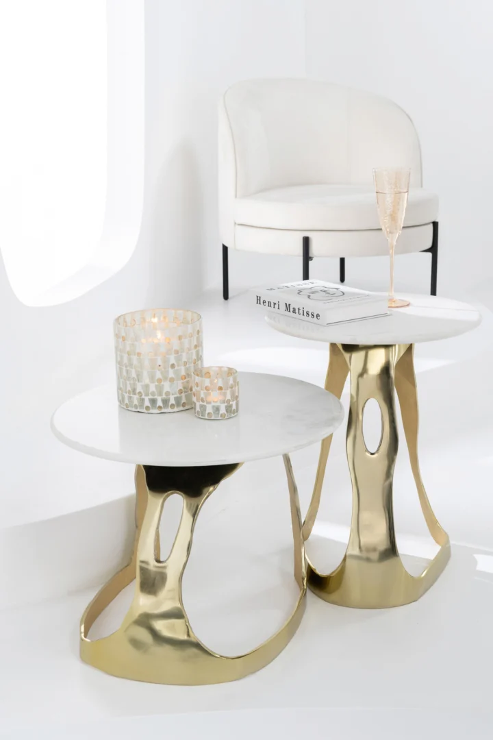 Twee aluminium tafeltjes van Bijzettafel Goud, Wit 51 x 38 CM staan naast een moderne witte fauteuil. Hierop staan twee glazen kandelaars, een boek en een champagneglas. De ruimte is licht en minimaal.