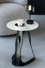 De Bijzettafel Zwart, Wit 41 x 48 CM, gemaakt van aluminium met een wit blad en zwart onderstel, bevat een koffiekopje en een kleine zwarte lamp naast een grijze stoel met structuur op een gladde lichtgrijze vloer.