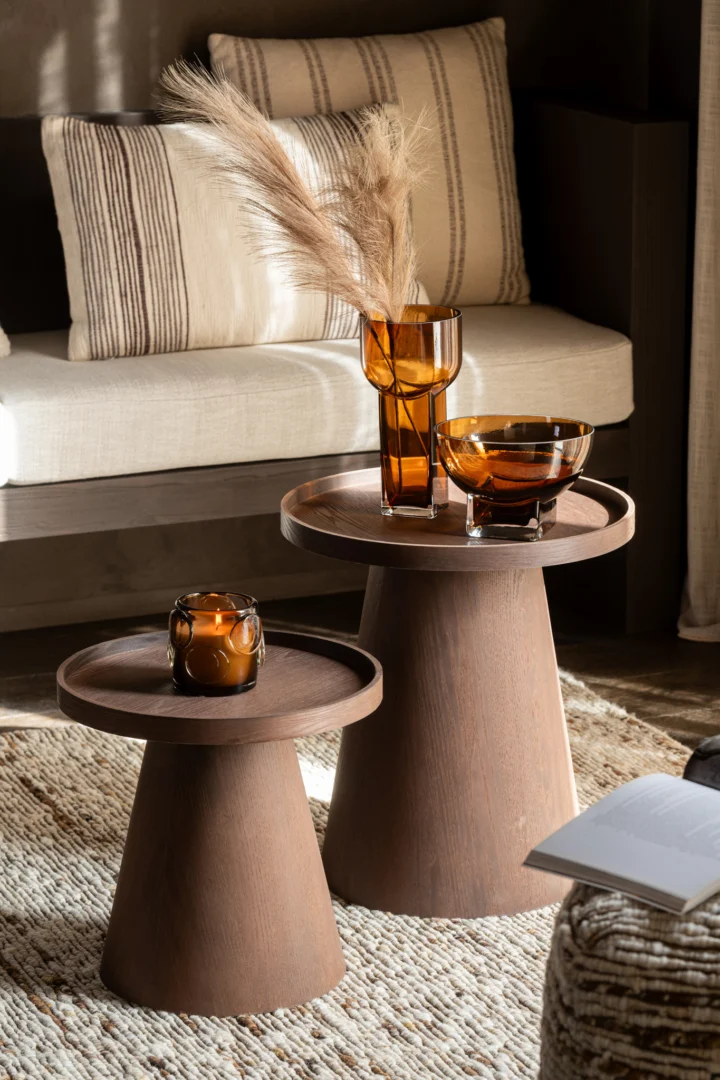 Een Bijzettafel 45 x 45 CM Rond Fineer toont amberkleurig glaswerk, een kaars en een glazen vaas met pampagras. Daarachter staat een beige bank met gestreepte kussens op een vloerkleed met textuur, naast een open boek.
