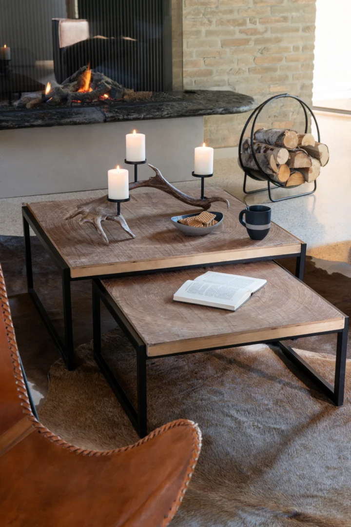 In een gezellige woonkamer staat de salontafel Zwart, Roest 80 x 63 CM Ijzer met kaarsen, een mok, een open boek, een gewei en snacks. Brandhout ligt opgestapeld bij een brandende open haard; een leren stoel verschijnt op de voorgrond.