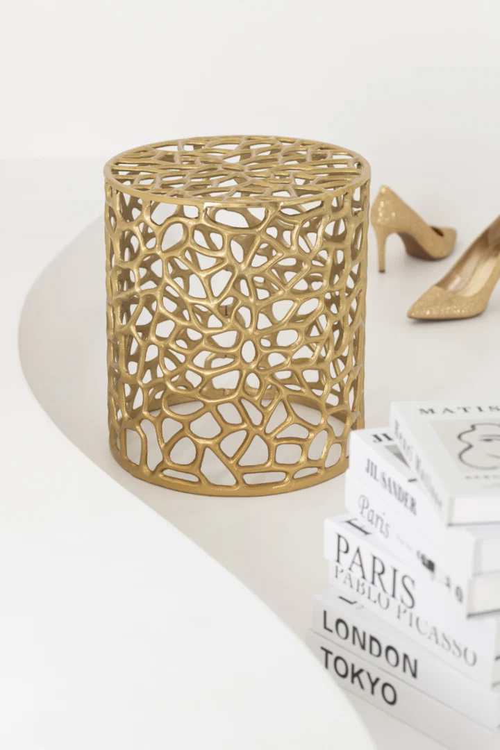 De S/2 Bijzettafel Aluminium Goudkleurig, een gouden opengewerkt metalen bijzettafeltje met een abstract patroon, staat op een wit oppervlak naast een paar gouden schoenen met hoge hakken en een stapel mode- en kunstboeken.