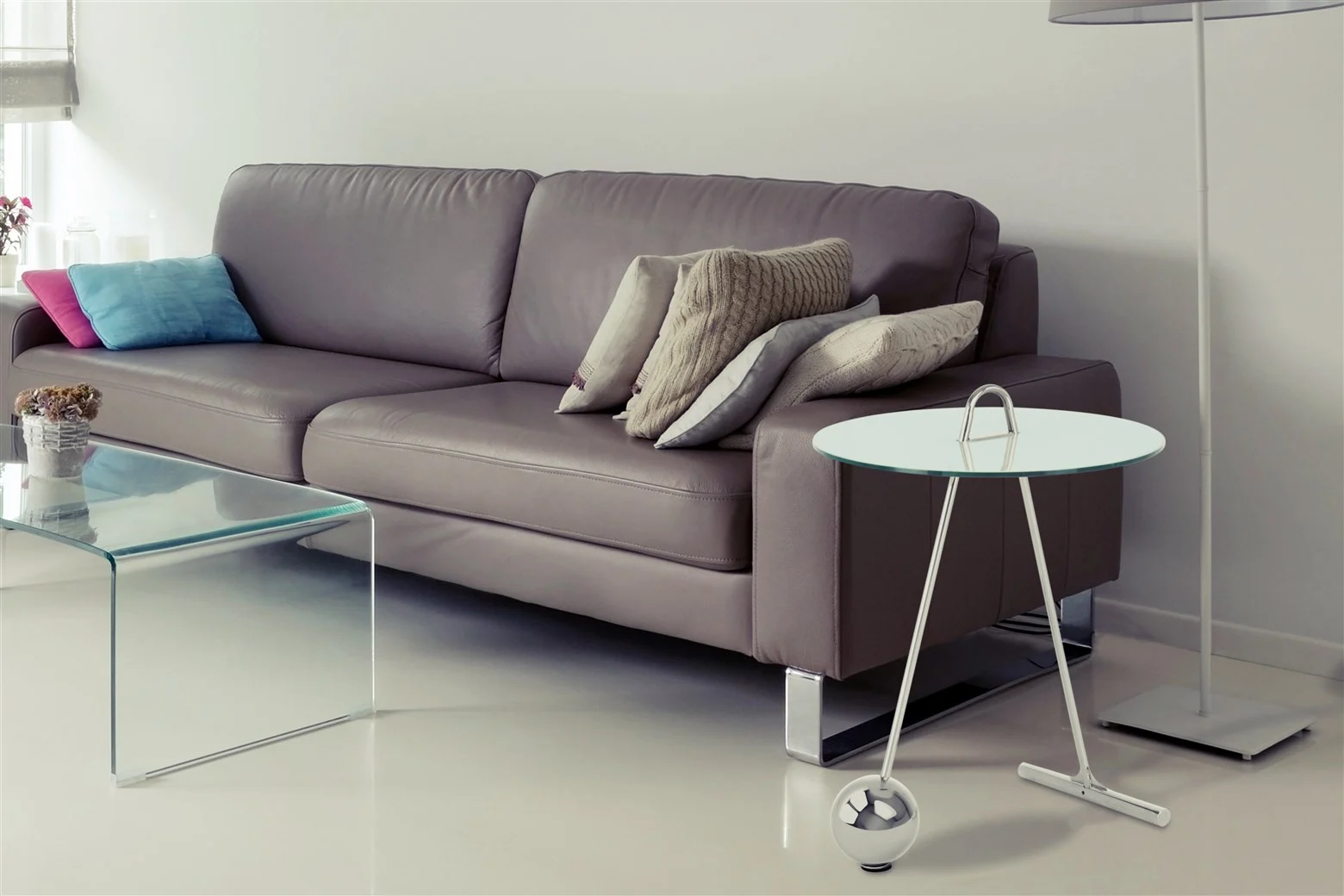 Een moderne woonkamer met een grijze leren bank, kussens, een glazen salontafel, een Bijzettafel Zilver, Wit Staal Compact, een lamp en een neutrale inrichting. De ruimte is licht en minimalistisch.