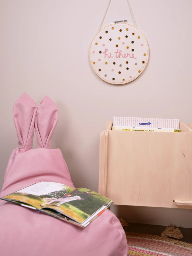 Een gezellig voorleeshoekje voor kinderen met de Zitzak Kind Kunstleer Pink bunny-ear zitzak, een opengeslagen prentenboek, een houten boekenhouder en een wandhanger met de tekst "hoi daar" in roze met gouden stippen.