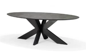 De Schakel Eettafel 220X100 Ovaal Keramiek + Stellar heeft een modern ovaal ontwerp met een donker, steenachtig keramisch blad en een opvallend zwart geometrisch onderstel van elkaar kruisende schuine poten, gepresenteerd op een effen witte achtergrond.