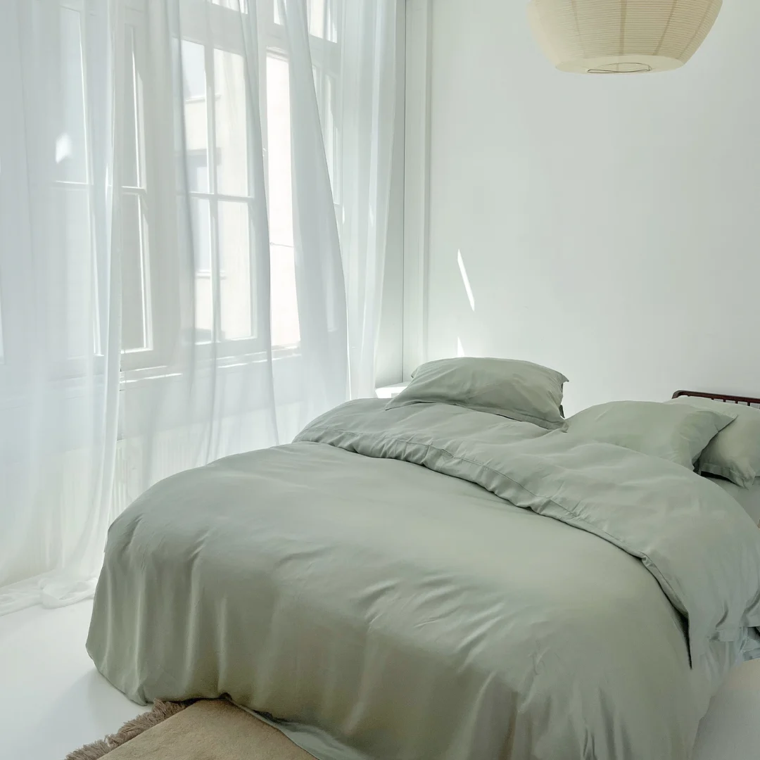 Een netjes opgemaakt bed met de Dekbedovertrek 140x220 Luxe uitstraling in lichtgrijs staat in een lichte, minimalistische kamer met transparante witte gordijnen en grote ramen. Een beige vloerkleed en een ronde papieren lantaarn dragen bij aan de serene stijl.