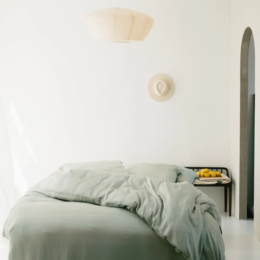 Een minimale slaapkamer met de Dekbedovertrek 140x220 Luxe uitstraling in lichtgroen op het bed, een tafeltje met citroenen en een kopje, een wandhoed en een geweven hanglamp, allemaal tegen witte muren die baden in zacht natuurlijk licht.