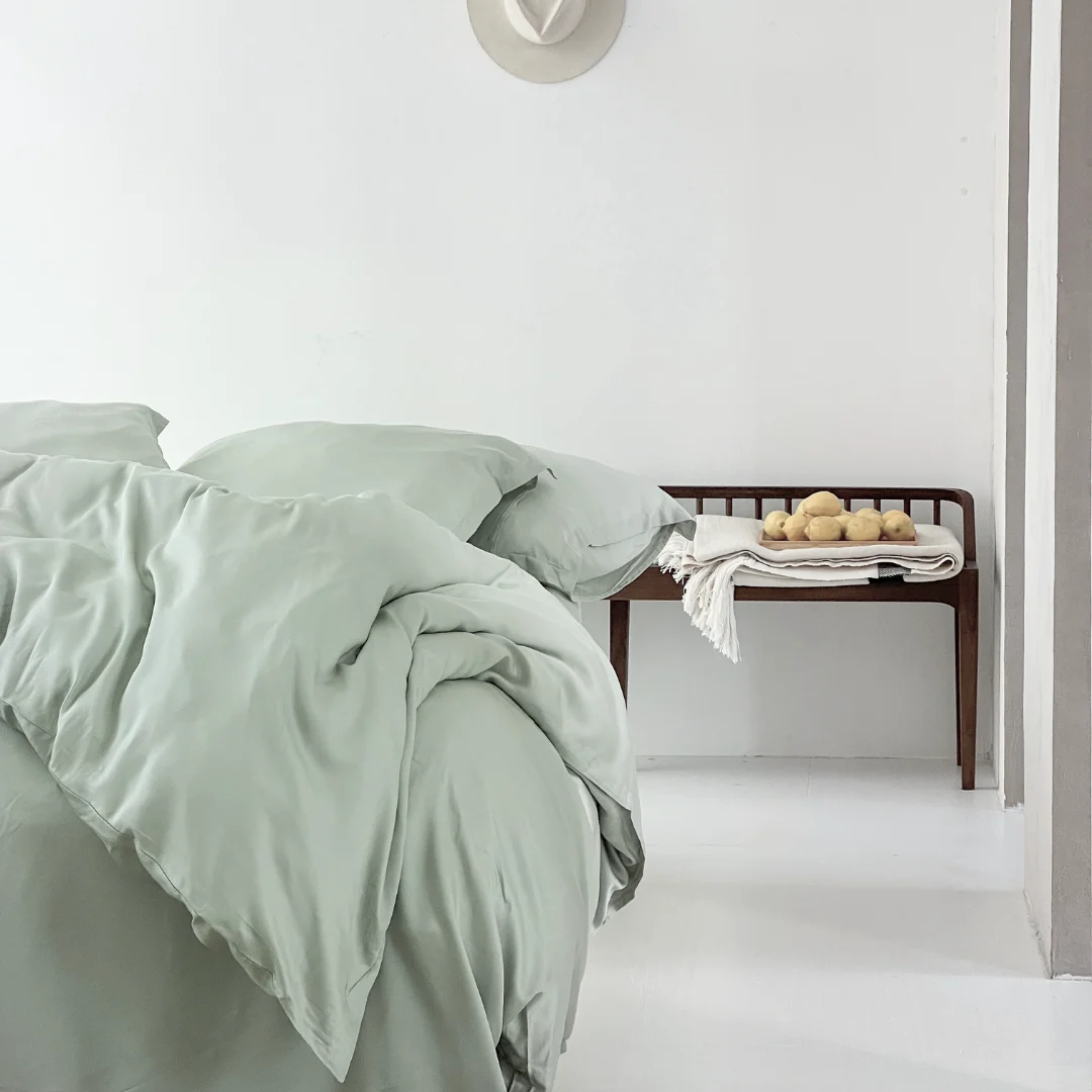 Lichtgroen hoeslaken van Tencel Lyocell opgemaakt op een bed in een witte kamer. Een houten bankje met fruit op de achtergrond. Zachte belichting.