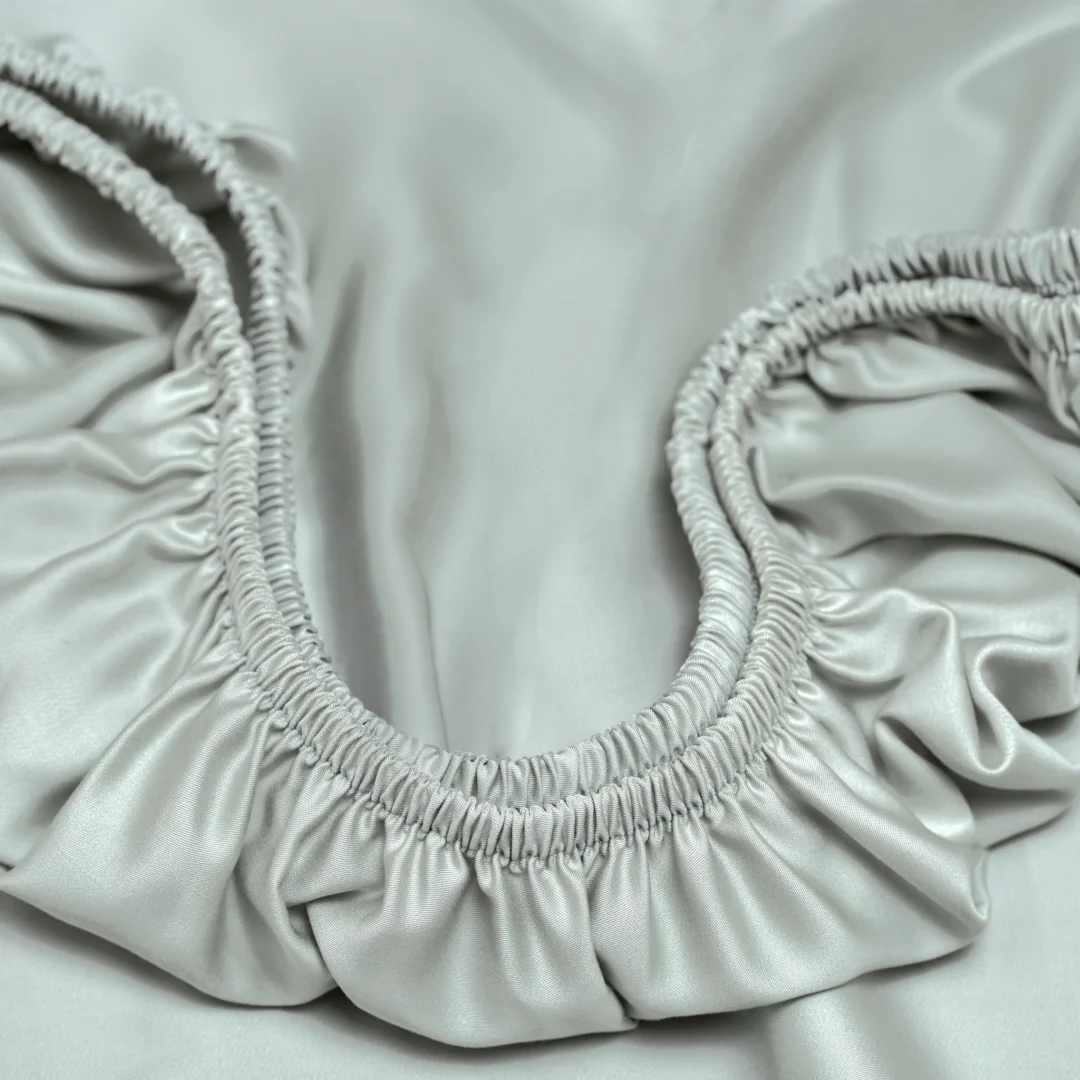 Close-up van een zacht, grijs hoeslaken met elastiek. De stof heeft een subtiele glans en valt in plooien. Zachte belichting.