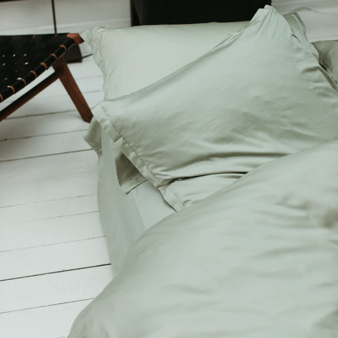 Detailopname van een bed met een zacht, groen hoeslaken. Witte houten vloer en een stoel op de achtergrond. Zachte belichting.