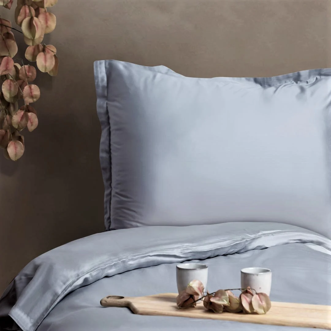 Een netjes opgemaakt bed met de Dekbedovertrek 200x200 Diervriendelijke keuze in lichtgrijs staat tegen een taupe muur. Op het bed staat een houten dienblad met twee kleine kopjes en gedroogde bloemen, terwijl aan de linkerkant van de afbeelding gedroogde bladeren hangen.