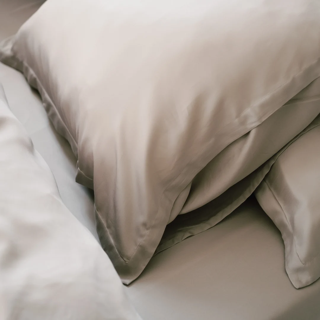 Close-up van een beige dekbedovertrek en kussensloop op een bed. Zachte belichting, gladde stof en subtiele stiksels zijn zichtbaar.