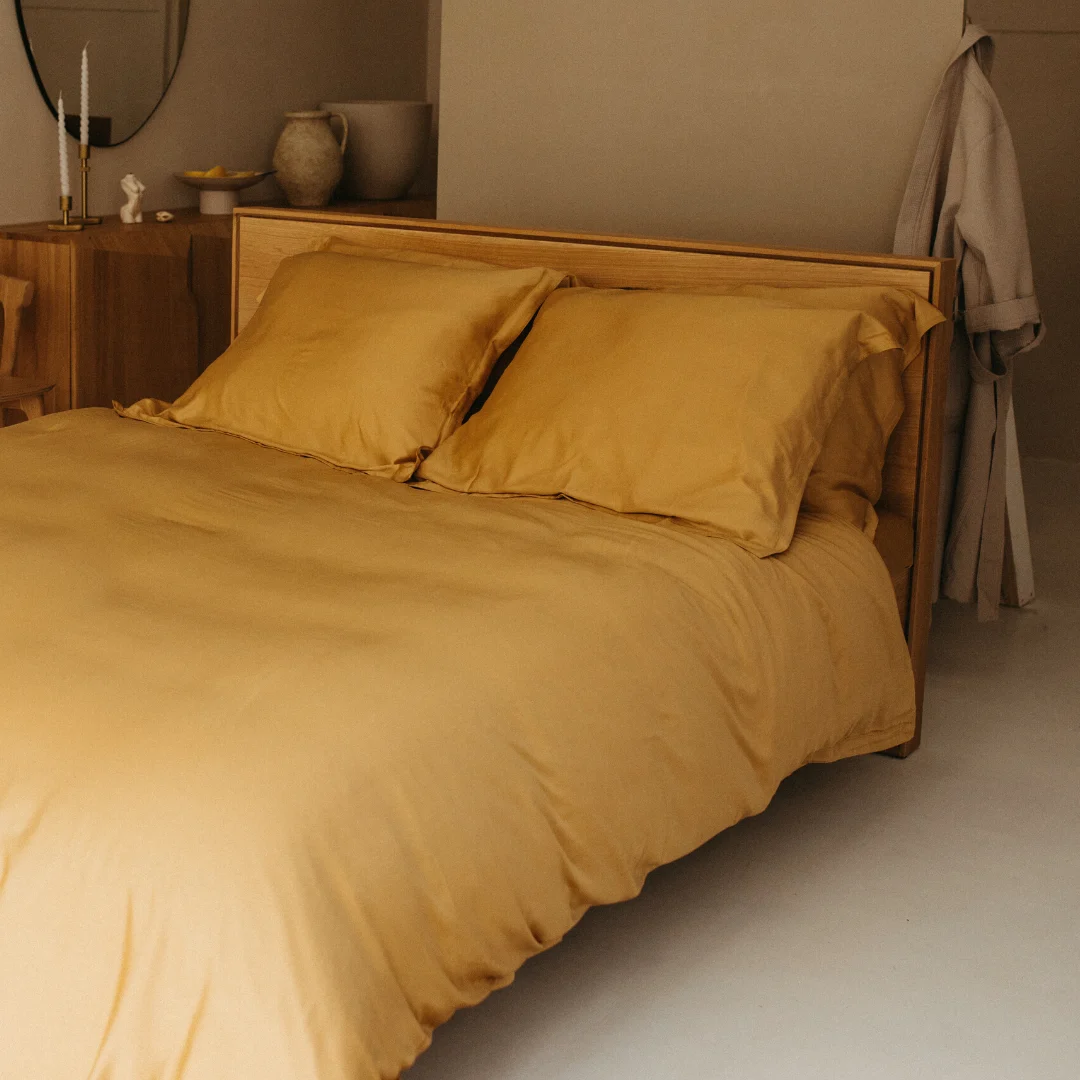 Overzicht van een bed met okergeel beddengoed en kussenslopen. Het bed heeft een houten frame en de kamer heeft een zachte, warme belichting.