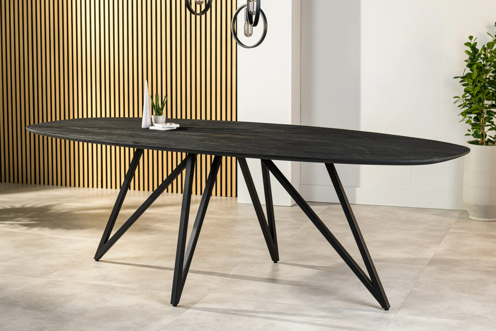 De Eettafel, 240x110 cm, B340 zwart is een moderne zwarte ovale eettafel met een geometrisch schuin onderstel, gestyled in een lichte kamer met een accentmuur van houten latten, een decoratieve plant en een minimalistisch tafeldecor.