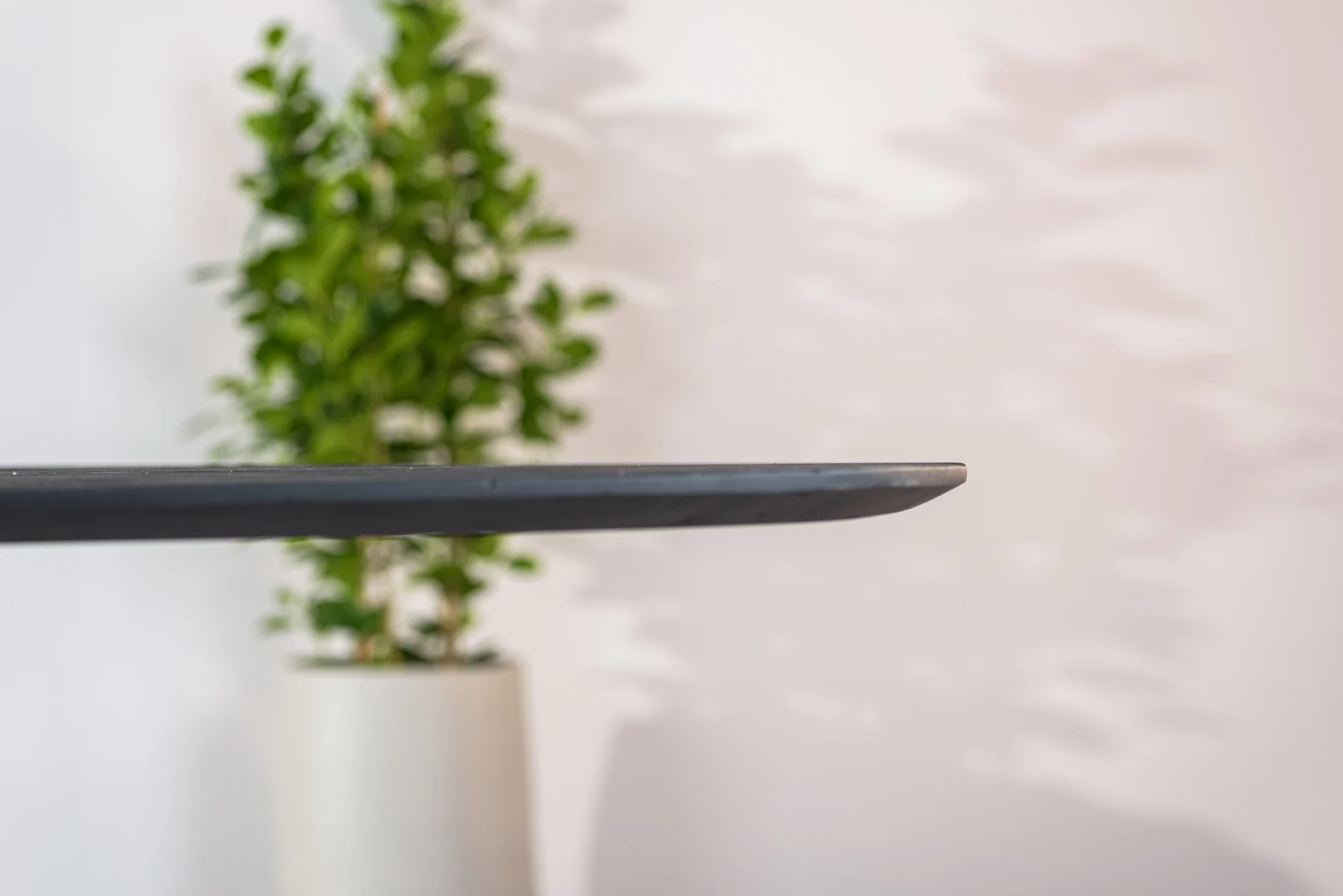 Een close-up van de rand van de Eettafel, 240x110 cm, B340 zwart laat het dunne, donkere tafelblad zien, met een vage groene potplant op de achtergrond tegen een witte muur.