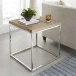 De Ace Teruggevonden Vuren en Gepolijst Roestvrijstalen Bijzettafel staat naast een lichte bank, met daarop boeken, een potplant en een glas. Vlakbij ligt een gestreept tapijt op de vloer.