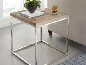 De Ace Teruggevonden Vuren en Gepolijst Roestvrijstalen Bijzettafel staat naast een lichte bank, met daarop boeken, een potplant en een glas. Vlakbij ligt een gestreept tapijt op de vloer.