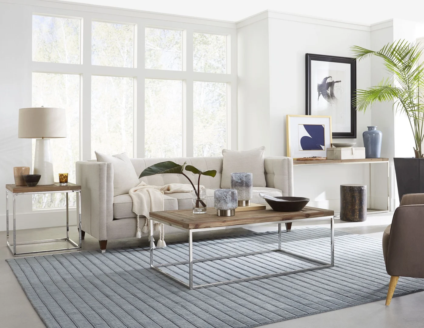 Een moderne woonkamer met een beige bank, houten salontafel, Ace Teruggevonden Vuren en Gepolijst Roestvrijstalen Bijzettafel met daarop een lamp, blauwgrijs vloerkleed, muurkunst, potplant en grote ramen die natuurlijk licht binnenlaten.