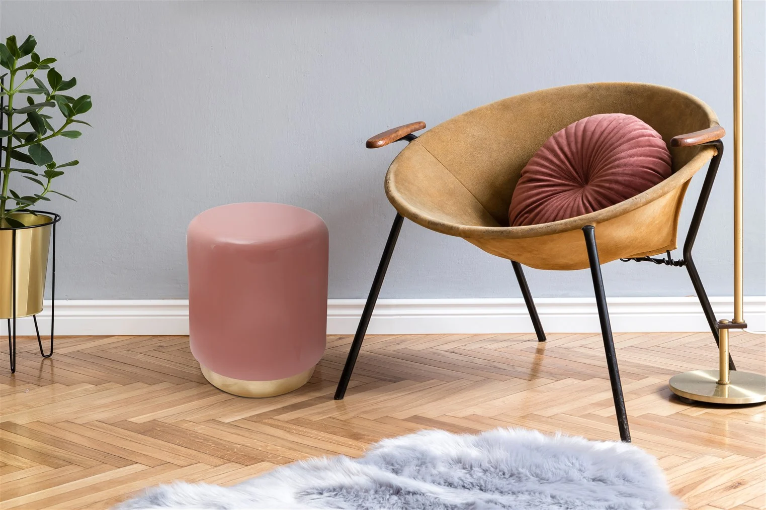 Een moderne kamer is voorzien van de Bijzettafel Roze Rond Ijzer Design, een bruine ronde stoel met een roze kussen, een roze voetenbankje, een gouden staande lamp, een plant in potten en een grijs vloerkleed van imitatiebont op een vloer van visgraathout.