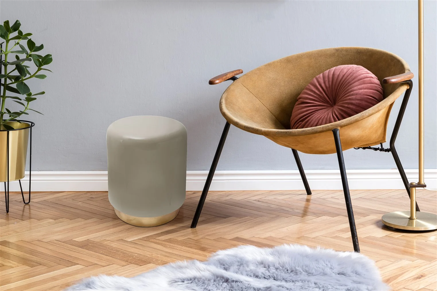 Een gezellig hoekje met een bruine moderne stoel, een rond roze kussen, een Bijzettafel Taupe Ijzer Chique, een potplant en een donzig vloerkleed op een visgraat houten vloer tegen een lichtgrijze muur.