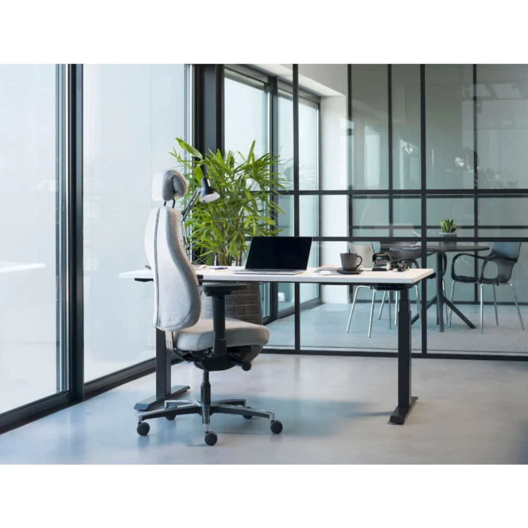 Modern kantoor met een wit bureau en de Bureaustoel Hoofdsteun, voorzien van een donkergrijs hoofdsteun, bij grote ramen. Op het bureau staan een laptop, lamp, beker en boek; op de achtergrond staan een groene plant en een vergaderruimte met glazen wanden.