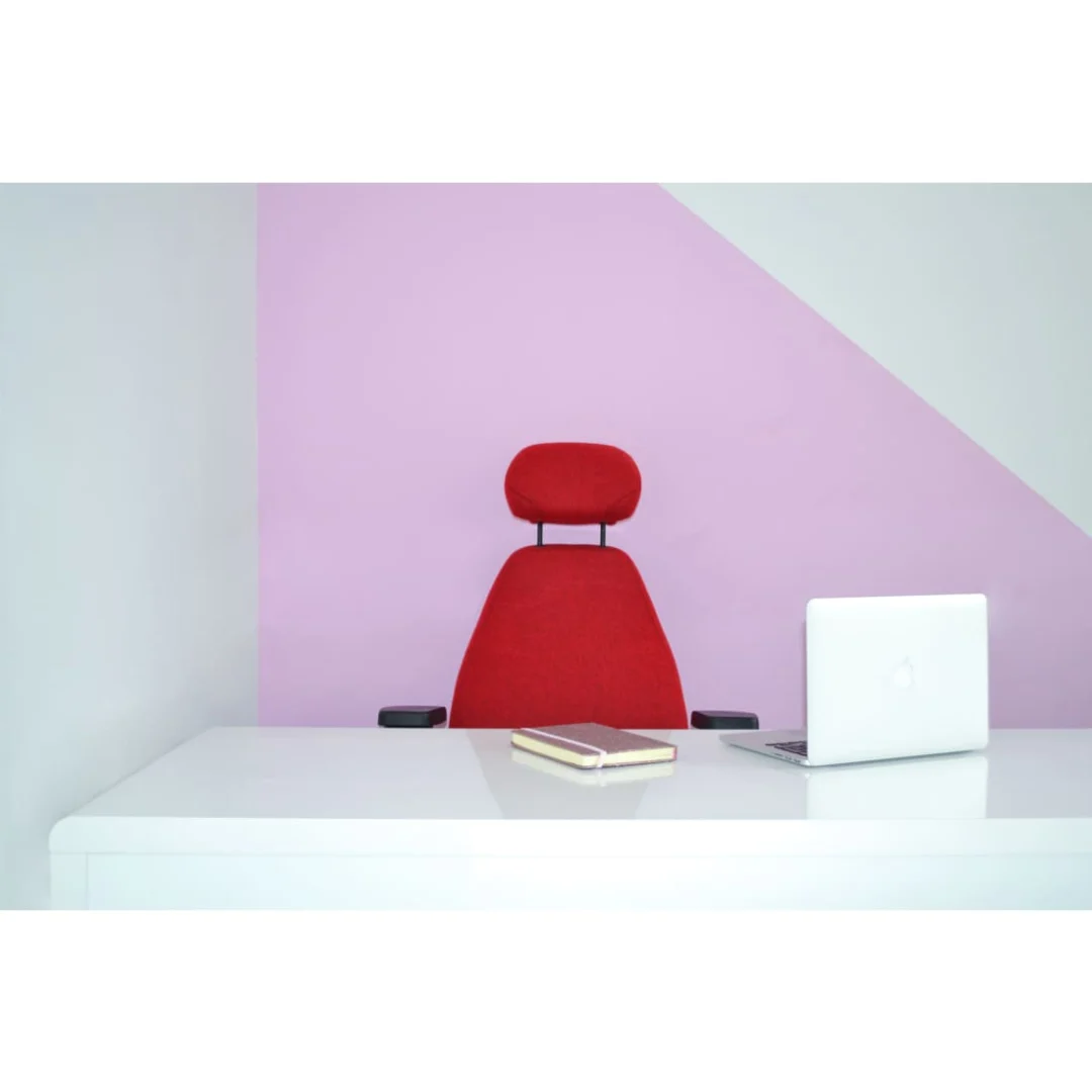 Een minimale kantooropstelling met een wit bureau, een Bureaustoel Hoofdsteun met daarachter een donkergrijs hoofdsteun, plus een gesloten zilveren laptop en twee notebooks, tegen een lichtroze en witte muur.