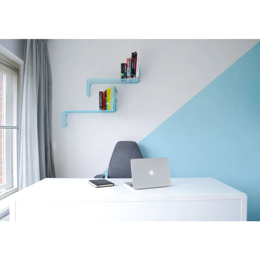 Een minimalistisch kantoor met een wit bureau, laptop, notitieblok en een Bureaustoel Hoofdsteun met donkergrijs hoofdsteun. Er zijn twee blauwe wandplanken met boeken, een geometrische witte en lichtblauwe muur en een raam met grijze gordijnen.