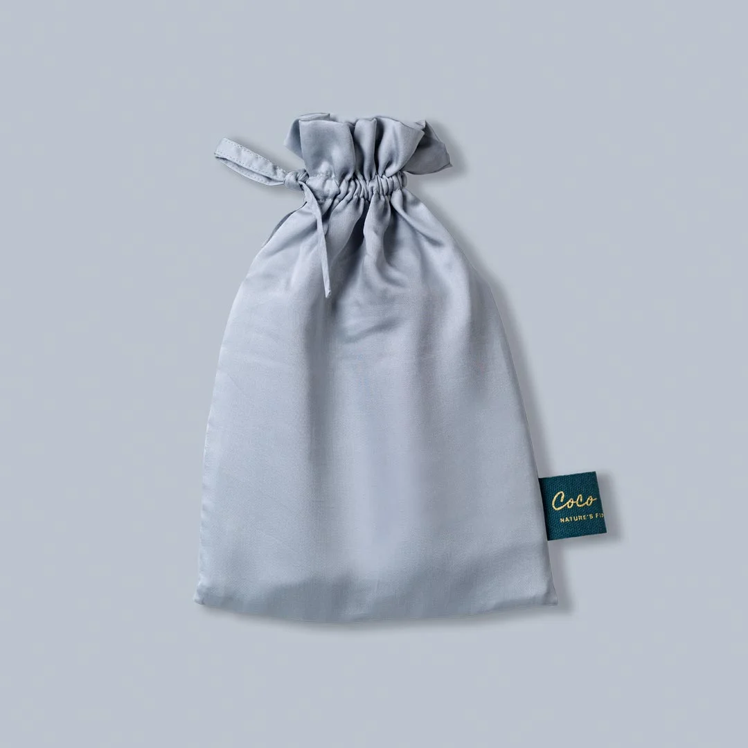 Lichtgrijze, satijnen zak met trekkoord, van bovenaf gefotografeerd. Label met gouden letters rechtsonder. Zachte belichting. Kussensloop.