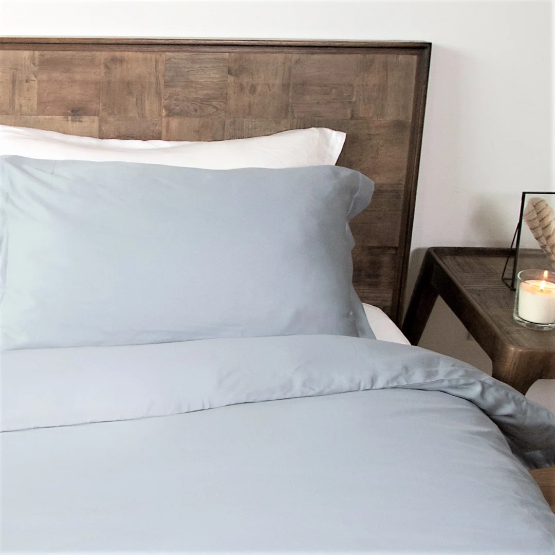 Lichtblauwe Tencel kussensloop opgemaakt op bed met houten hoofdeinde. Zachte belichting, rustige sfeer. Nachtkastje met kaars op de achtergrond.