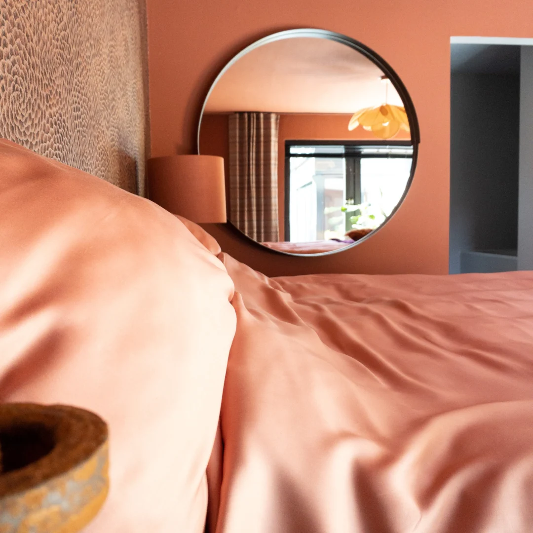 Detailopname van een roze dekbedovertrek opgemaakt bed. Ronde spiegel aan de wand reflecteert een lichte kamer. Zachte, warme belichting.