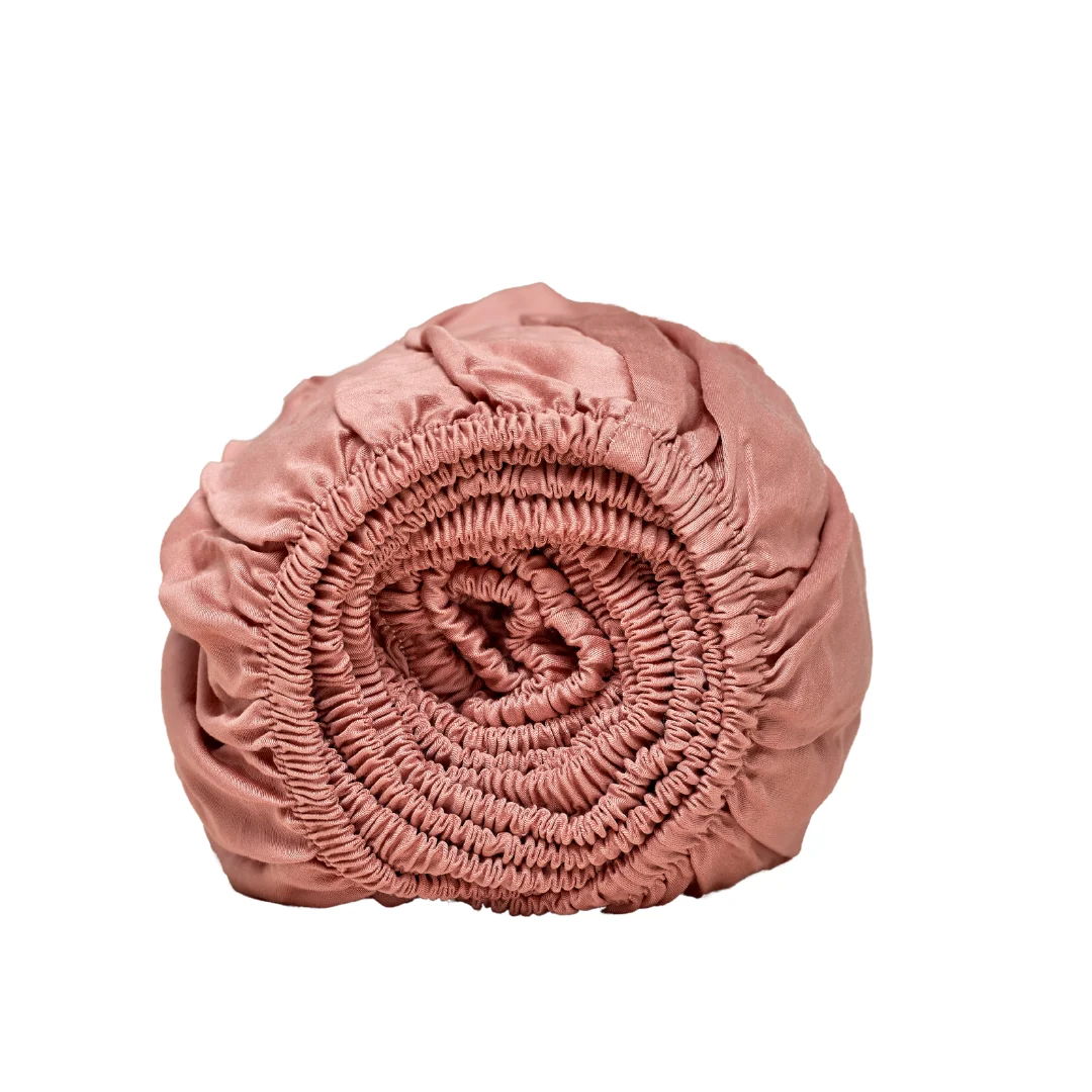 Oprolbaar, terracotta roze hoeslaken van Tencel satijn. Elastische randen zijn zichtbaar. Zachte stof en neutrale achtergrond.