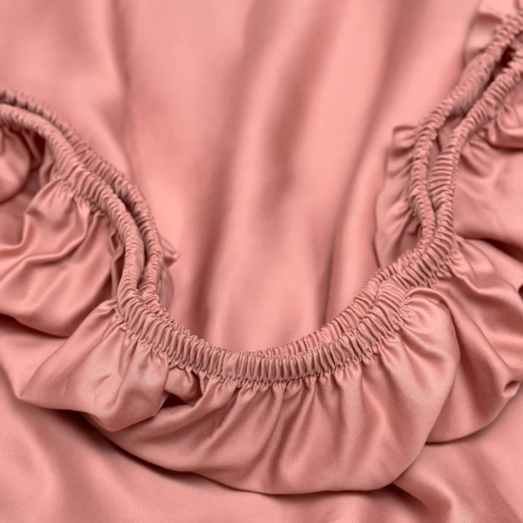 Close-up van een terracotta roze hoeslaken met elastiek, gemaakt van satijn Tencel. Zachte belichting benadrukt de luxe textuur.