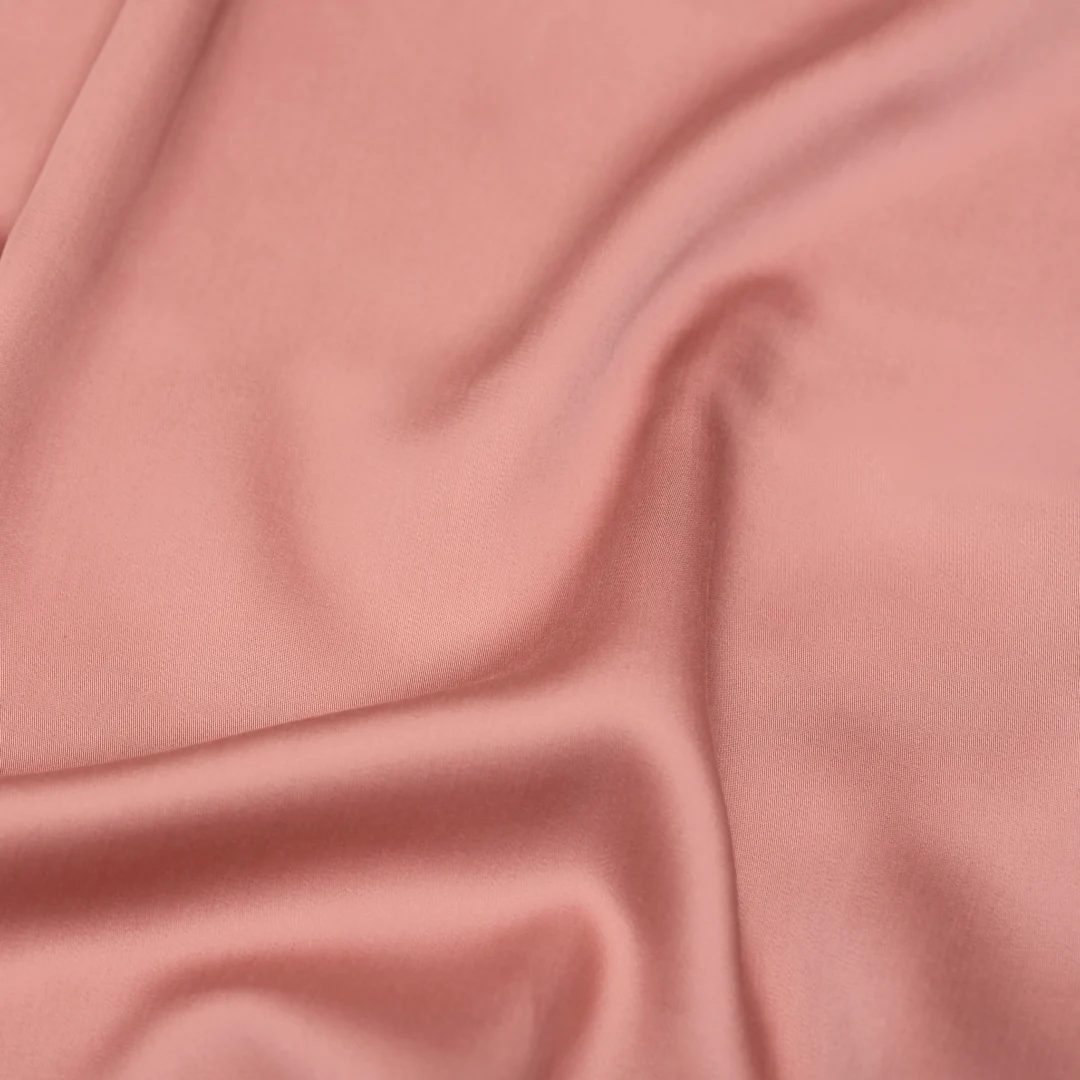 Close-up van een roze satijnen hoeslaken, met zachte plooien en een subtiele glans. Het luxe tencel materiaal valt soepel in het licht.