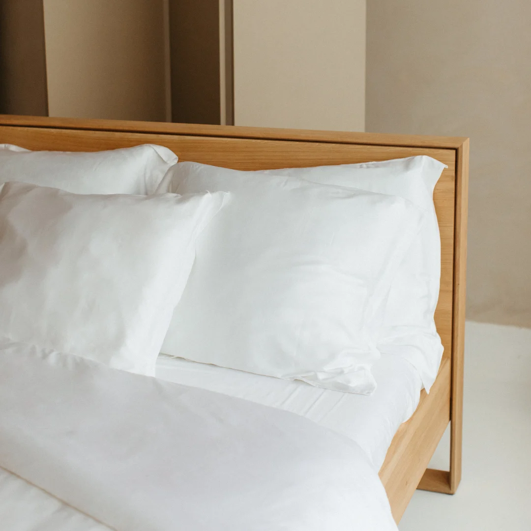 Detailopname van een houten bed met wit satijnen beddengoed en kussens. Zachte, natuurlijke belichting benadrukt de textuur van het hoeslaken.