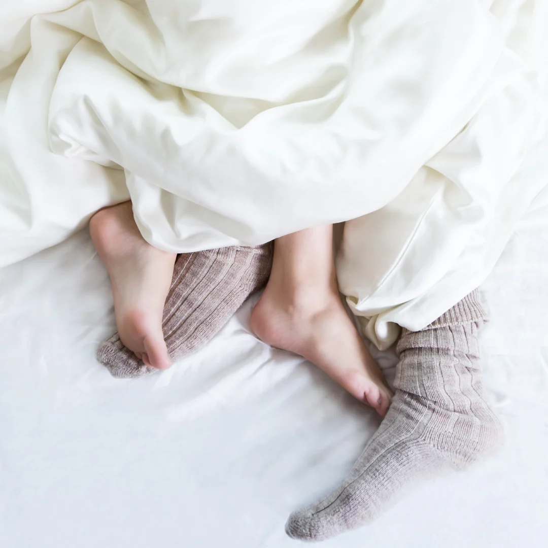 Twee paar voeten rusten op een bed met de Dekbedovertrek 200x220 Makkelijk gecombineerd; het ene paar is bloot, het andere in gezellige grijze gebreide sokken, beide onder een zachte witte deken.