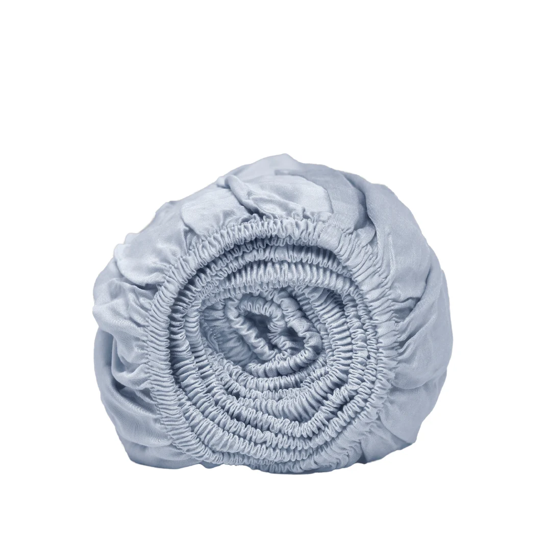 Oprolbaar lichtblauw hoeslaken met elastiek, satijnzacht. Zachte kleuren en gladde textuur. Ideaal voor een luxe slaapkamer. Witte achtergrond.