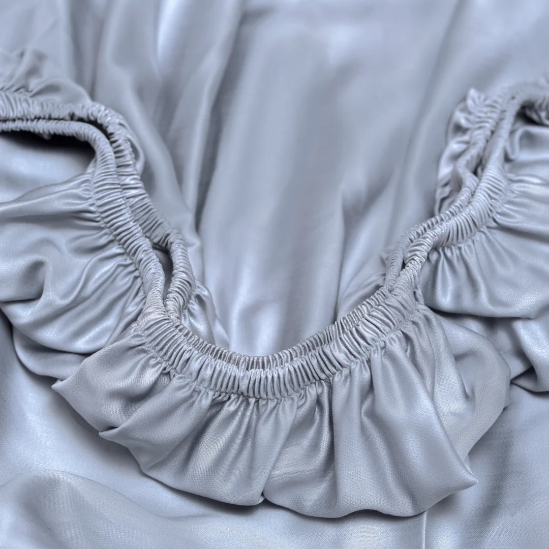 Close-up van een satijnzacht, grijs hoeslaken met elastiek. Zachte belichting accentueert de luxe textuur en de plooien van het Tencel lyocell.