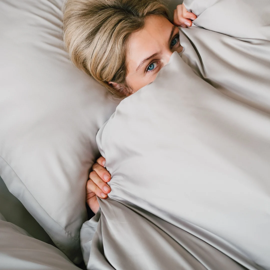 Een persoon met blond haar ligt in bed, grotendeels bedekt door het Dekbedovertrek 200x220 Vormvast, met alleen de ogen en handen die onder de lichtgrijze dekens vandaan komen.