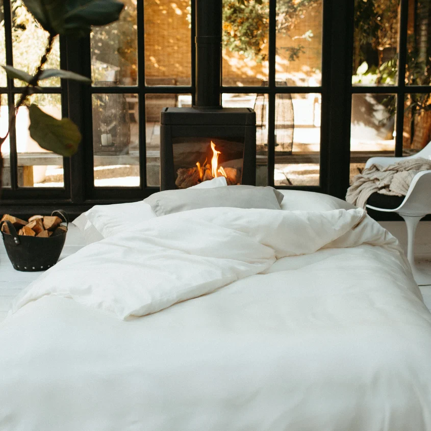 A Dekbedovertrek 200x220 Topkwaliteit textiel siert een knus bed voor een brandend haardvuur. Grote ramen bieden uitzicht op de zonnige tuin, terwijl een stoel en houtmand het warme, uitnodigende sfeertje compleet maken.