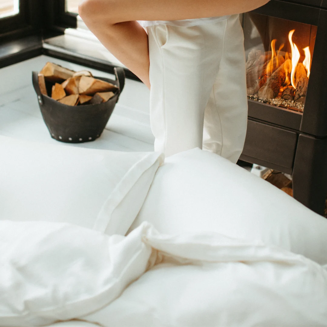 Een persoon in witte pyjama staat bij een bed met de Dekbedovertrek 260x220 Ademend comfort, naast een brandende open haard en een zwarte mand met brandhout, waardoor een gezellige, warme sfeer ontstaat.