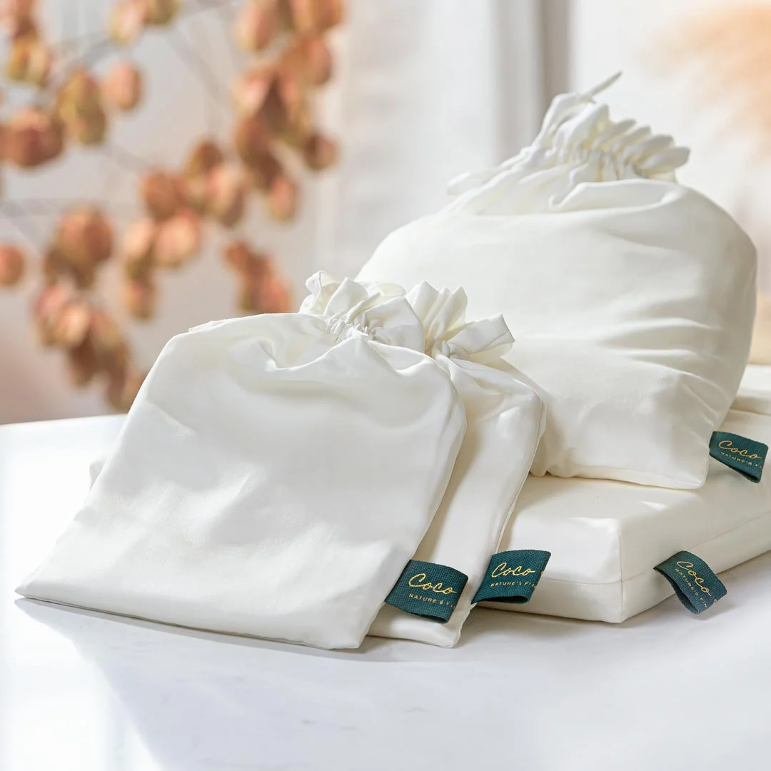 Drie Dekbedovertrek 260x220 Ademend comfortsets worden getoond op een wit oppervlak, elk met een witte stoffen zak, koordsluiting en groen "Coco"-label. Bladeren in warme tinten creëren een zacht vage achtergrond.